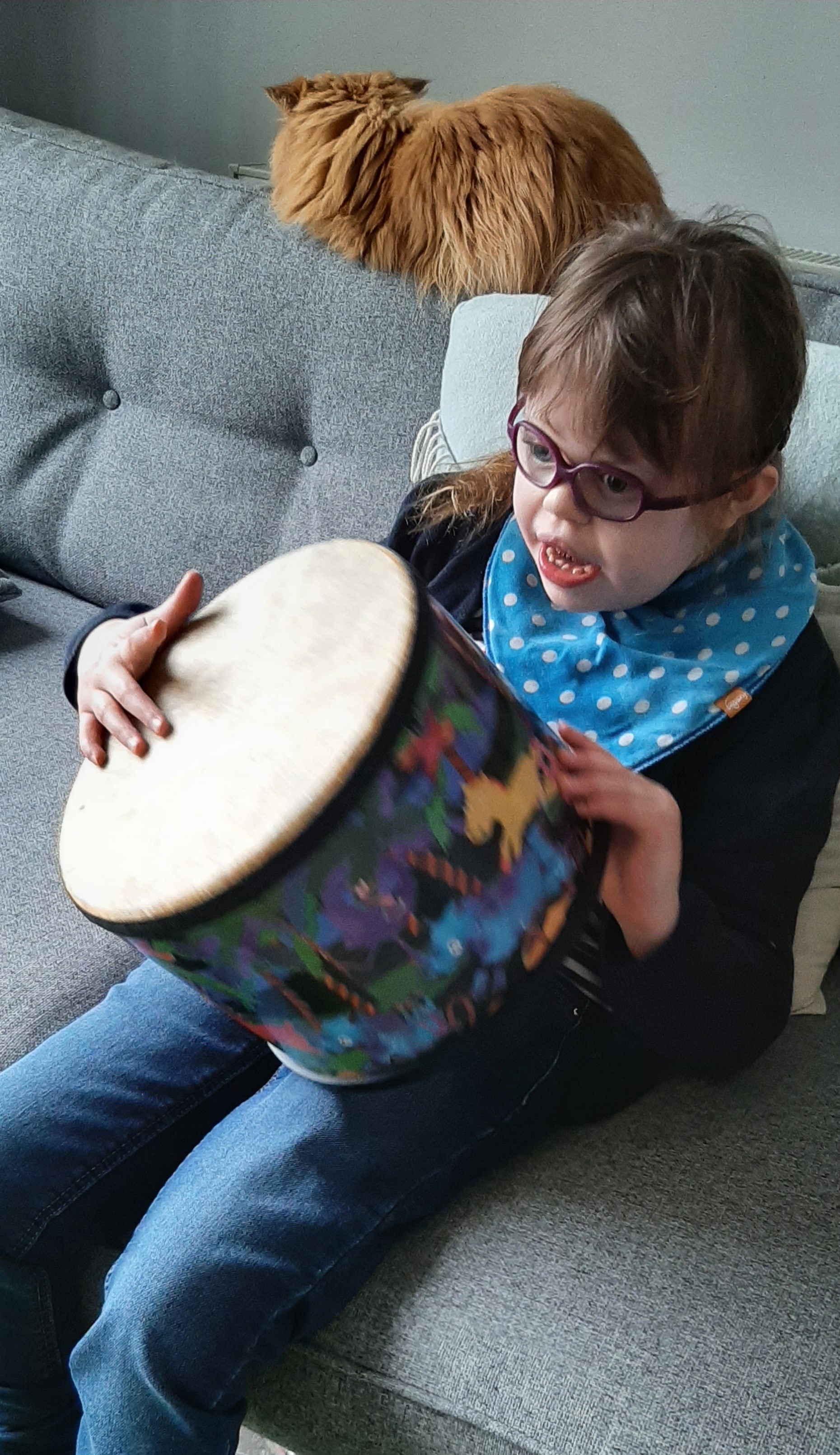 Lucy is sitting on her sofa, holding and playing her drum.